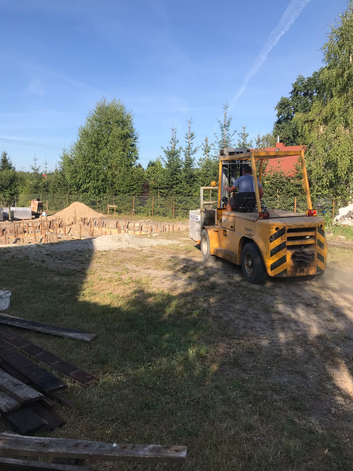 Żółty wózek widłowy Fenwick z operatorem na placu budowy, widoczne wykopane fundamenty z drewnianym szalunkiem i sterta piasku, w tle drzewa iglaste i ślad po samolocie na błękitnym niebie.