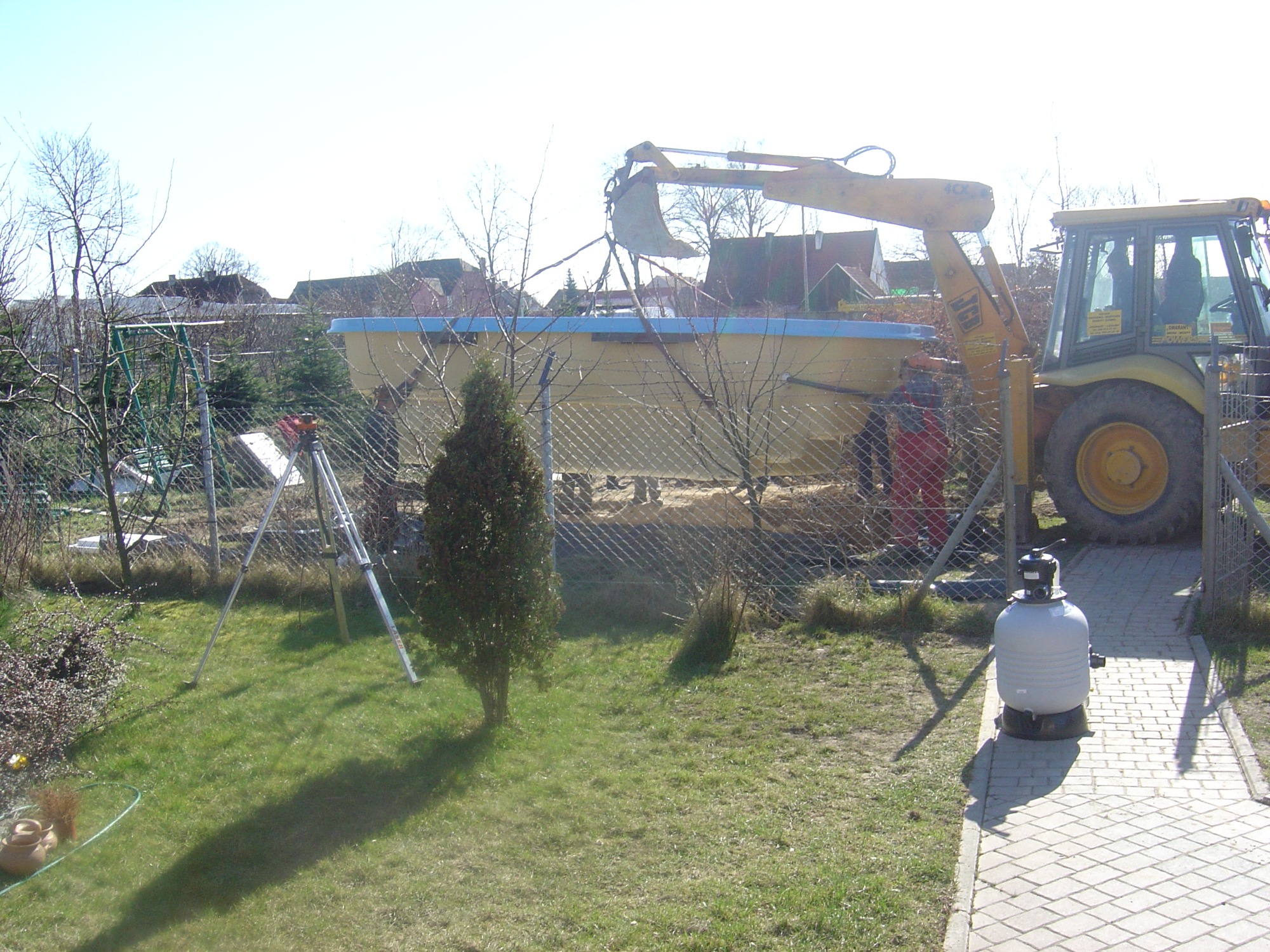 Żółta koparko-ładowarka podnosi jasny, pusty basen ogrodowy nad ogrodzeniem z siatki; w tle domy jednorodzinne, na pierwszym planie trawnik z małym drzewkiem i biały zbiornik filtrujący.