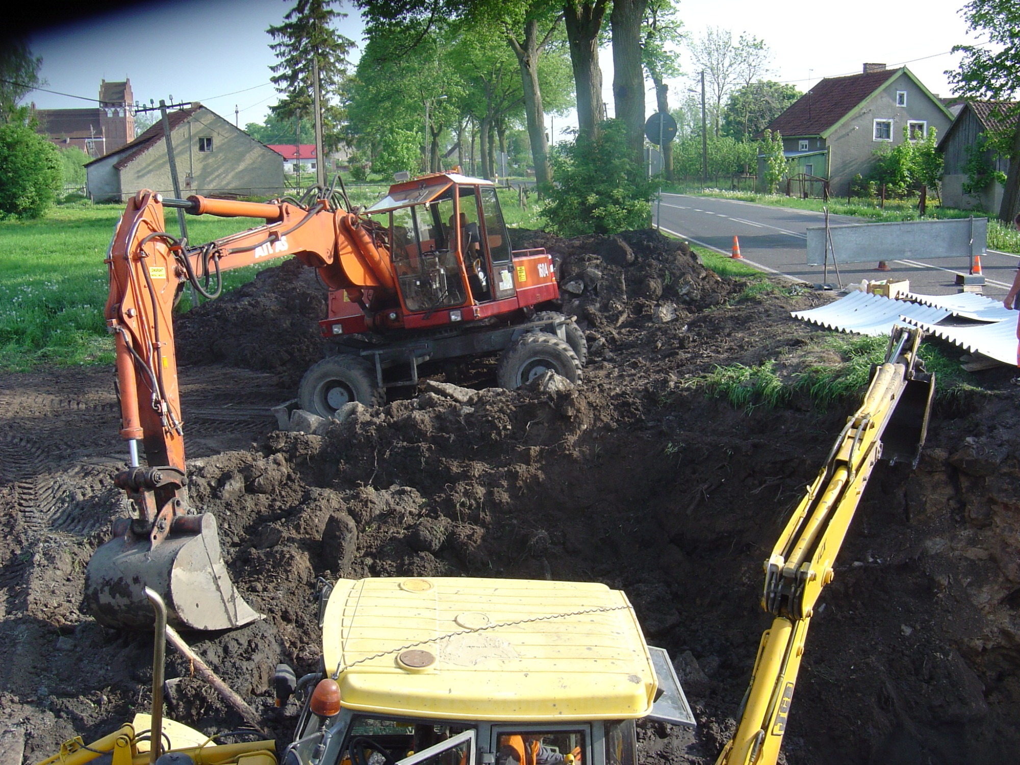 Dwa pracujące równocześnie, pomarańczowo-czerwone i żółte, ciężkie koparki na nierównym, błotnistym terenie z wykopem, w tle wiejskie zabudowania i droga zabezpieczona barierą.