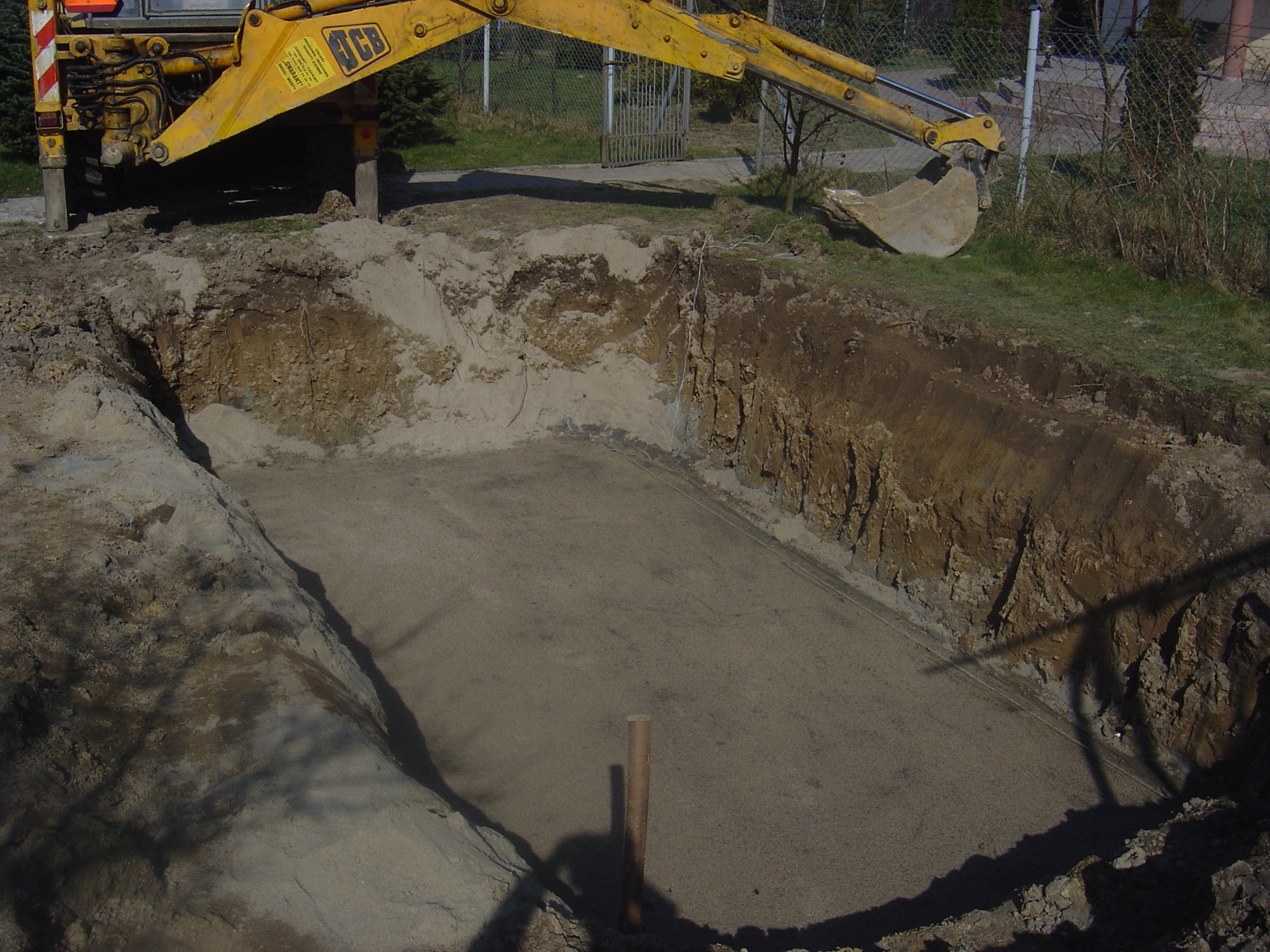 Żółta koparka JCB nad wykopem o prostokątnym kształcie z ubitym piaskowym dnem, widoczny drewniany palik w rogu, ziemia wokół wykopu.