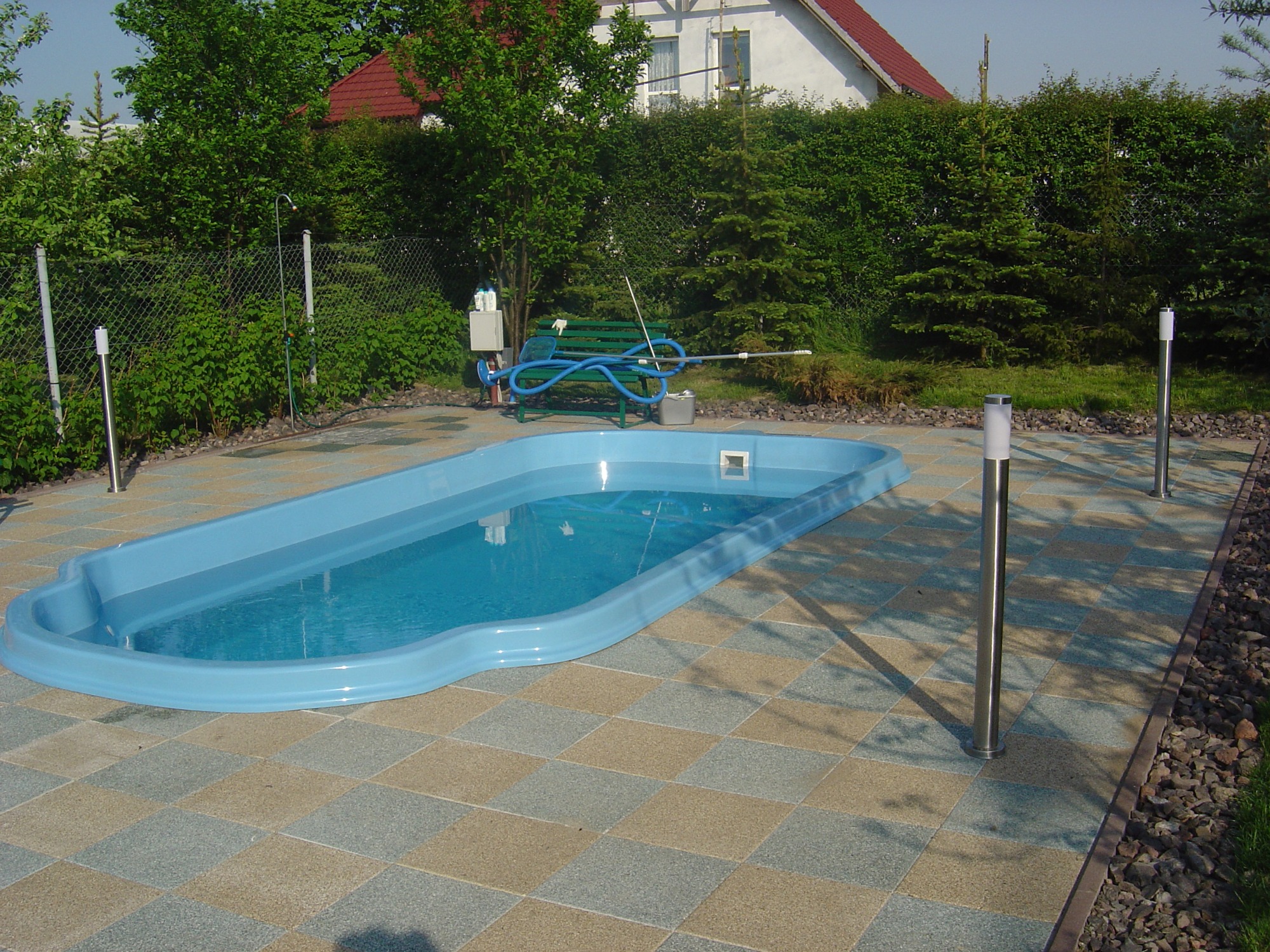 Niebieski basen ogrodowy otoczony kostką brukową w szachownicę, z widocznym sprzętem do czyszczenia, oświetleniem punktowym oraz zielenią w tle.
