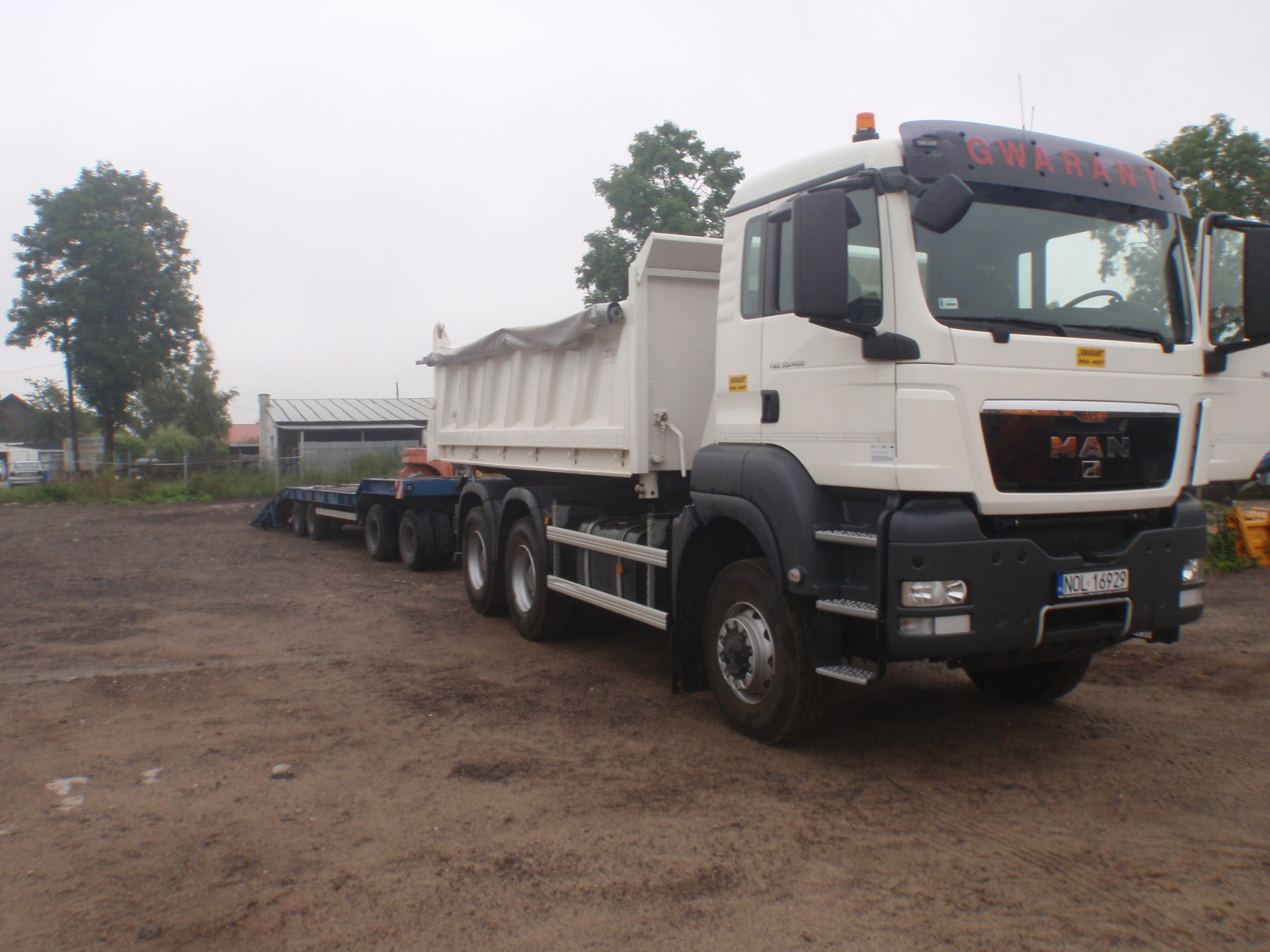 Ciężarówka MAN z białą zabudową skrzyniową i przyczepą niskopodwoziową na placu budowy, widoczne tablice rejestracyjne i napis 'GWARANT' na górnej krawędzi kabiny.