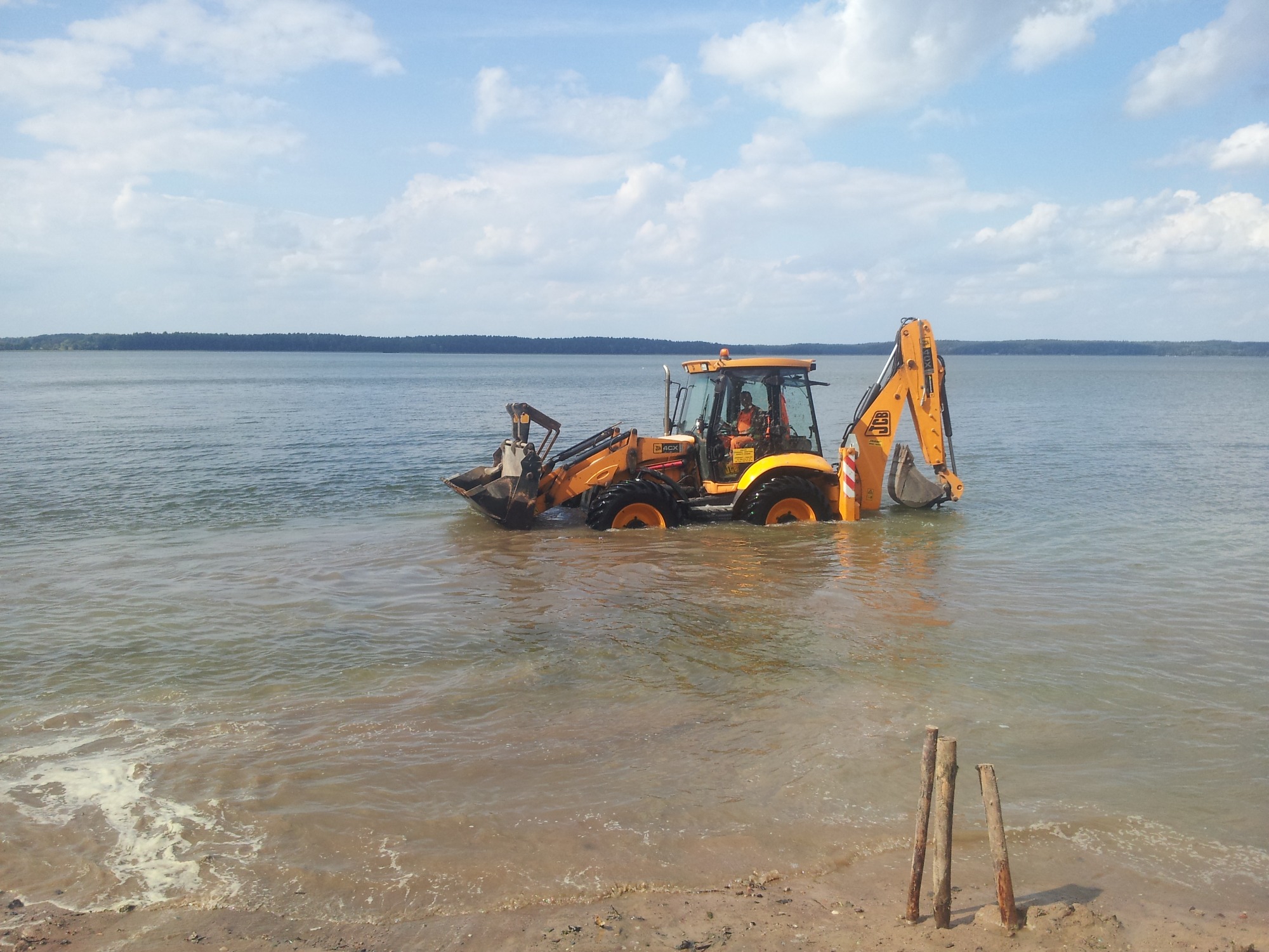 Żółta koparko-ładowarka JCB zanurzona w wodzie jeziora, z widocznym operatorem w kabinie, pracująca przy brzegu z wystającymi palikami w piasku.
