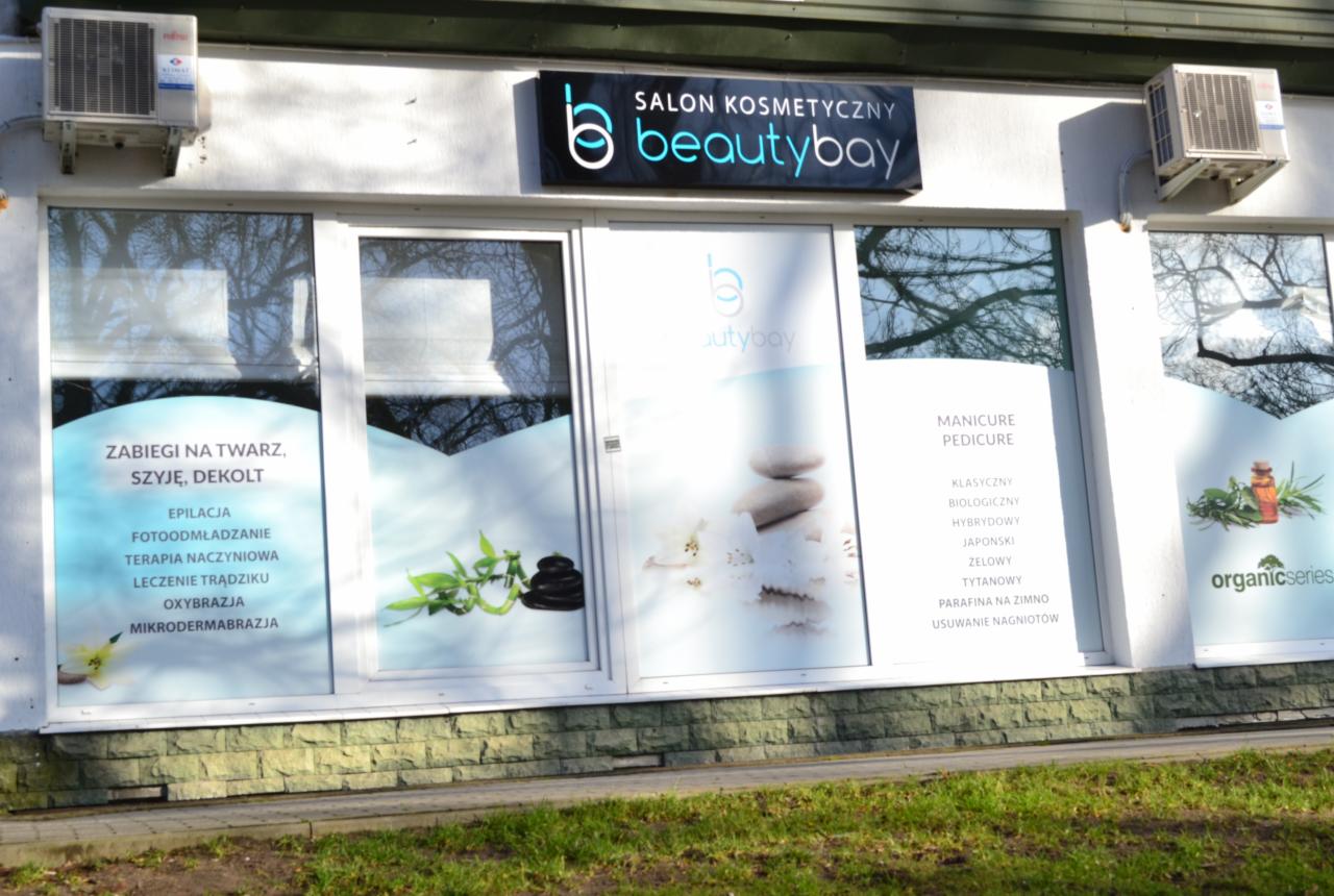 Fasada salonu kosmetycznego Beautybay z widocznymi naklejkami na oknach informującymi o ofercie: zabiegi na twarz, szyję i dekolt, manicure i pedicure oraz logo firmy na górze.