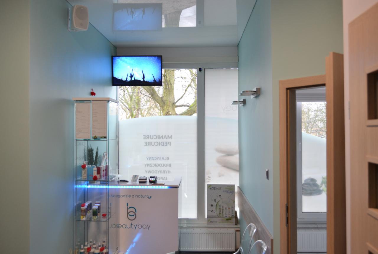 Wnętrze salonu kosmetycznego: recepcja z podświetlaną witryną, okno z napisem 'Manicure Pedicure', telewizor z motywem pustynnym, minimalistyczny design.
