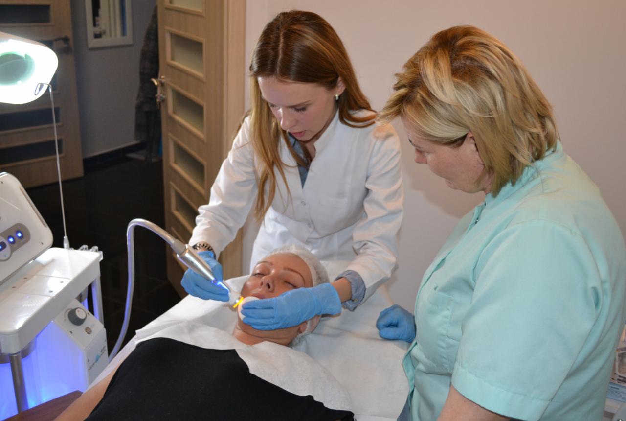 Kobieta poddawana zabiegowi na twarz z użyciem urządzenia emitującego niebieskie światło, wykonywanemu przez dwie kosmetyczki w rękawiczkach, w salonie kosmetycznym.