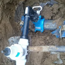 Podłączenie rur wodociągowych w wykopie ziemnym, widoczne złącza i rury z tworzywa sztucznego, niebieskie elementy mocujące.