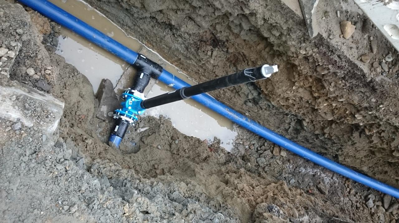 Niebieskie rury wodociągowe z zaworem i czarnym przyłączem ułożone w wykopie wypełnionym wodą i błotem.