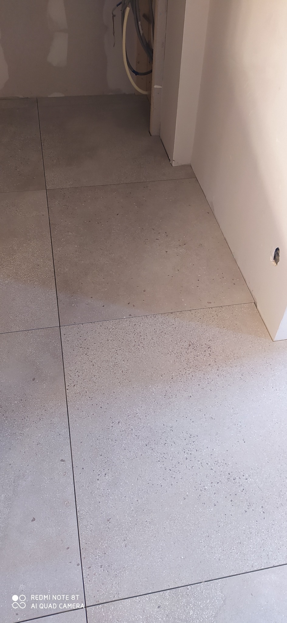 Ułożone szare płytki podłogowe z drobnym wzorem, widoczne rury instalacyjne przy ścianie w tle, ściany w trakcie wykańczania.