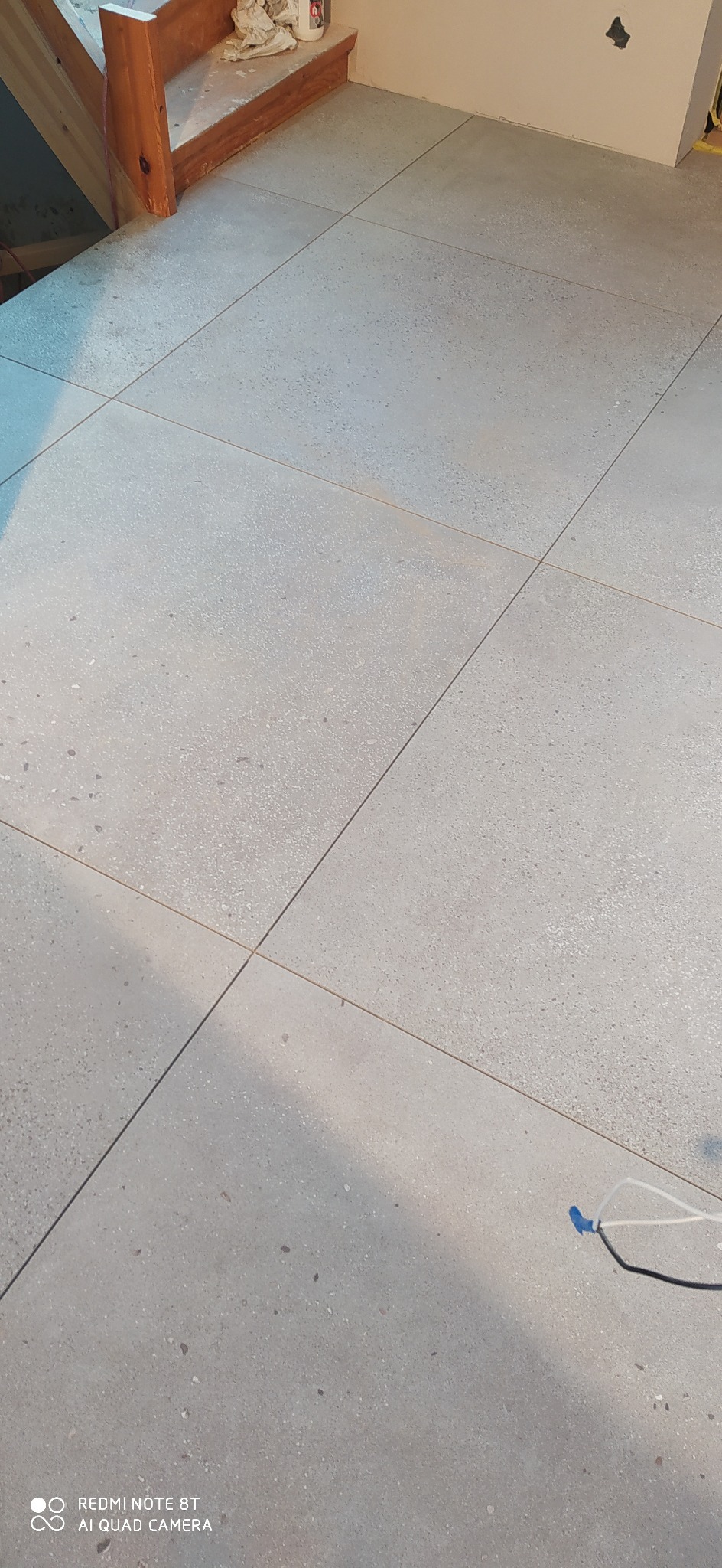 Ułożone, jasnoszare płytki podłogowe z delikatnym wzorem, widoczne fragmenty schodów i kabla w rogu.