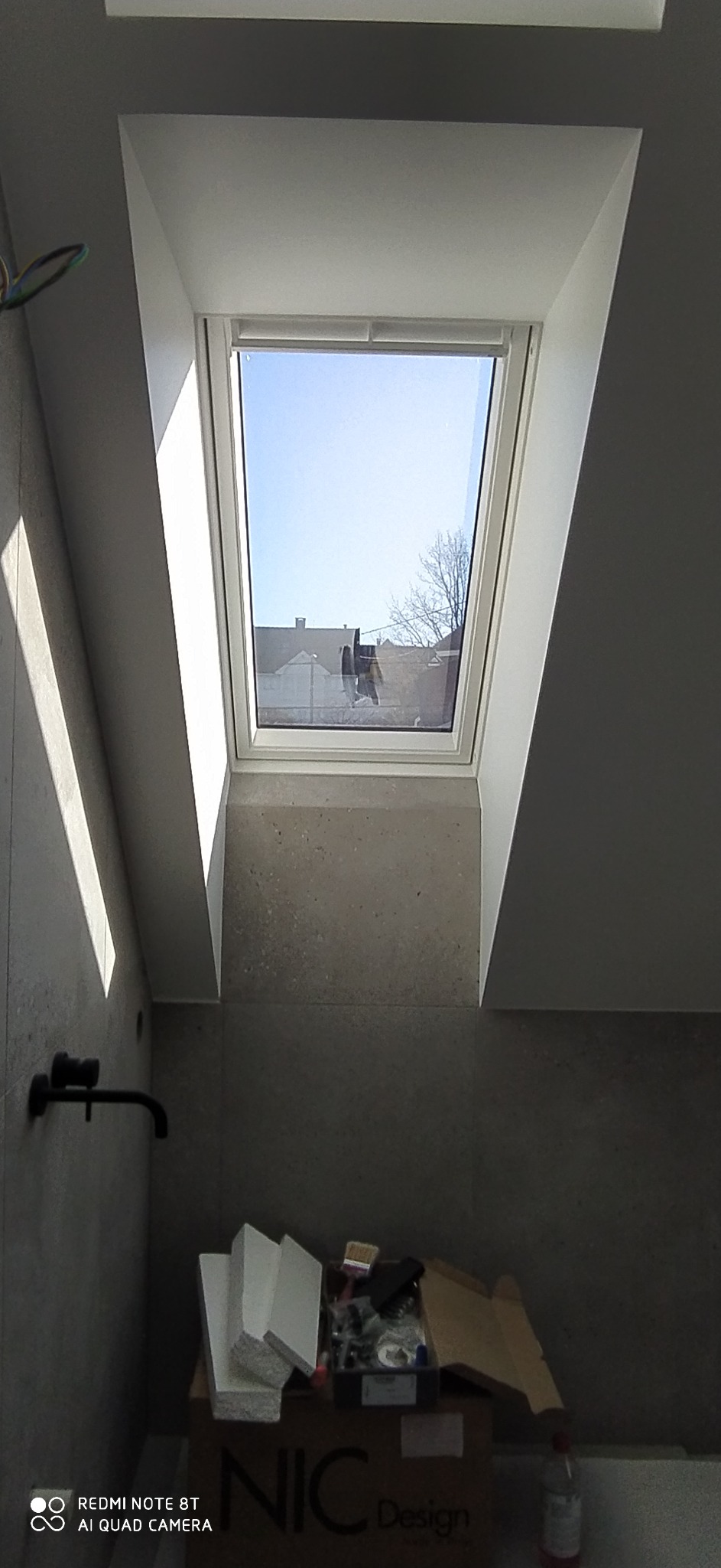 Poddasze łazienki z oknem dachowym, szare płytki na ścianach, czarna bateria umywalkowa i materiały budowlane w kartonie na podłodze.