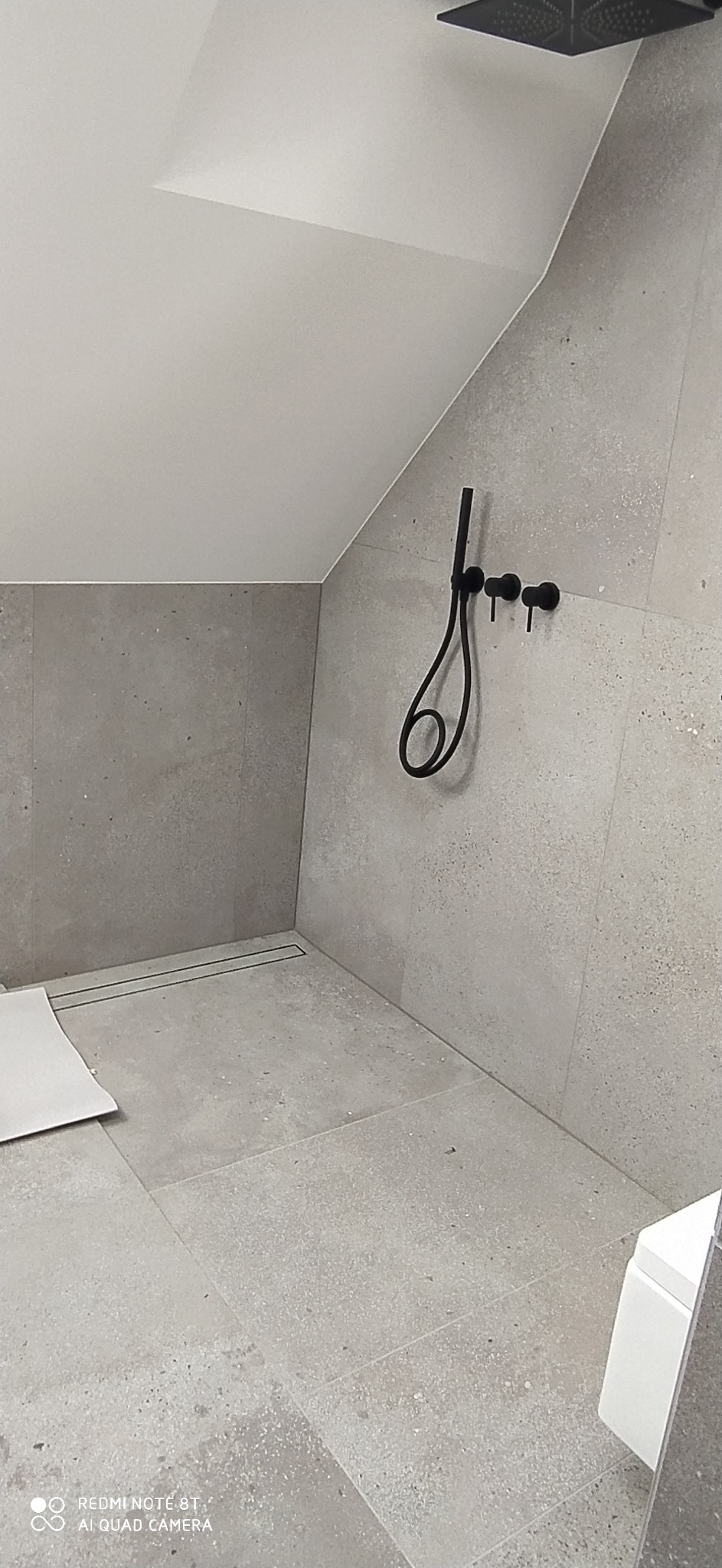 Wykończona łazienka na poddaszu z szarymi płytkami, czarną armaturą prysznicową i odpływem liniowym w podłodze, widoczne fragment toalety i niezabudowana przestrzeń.