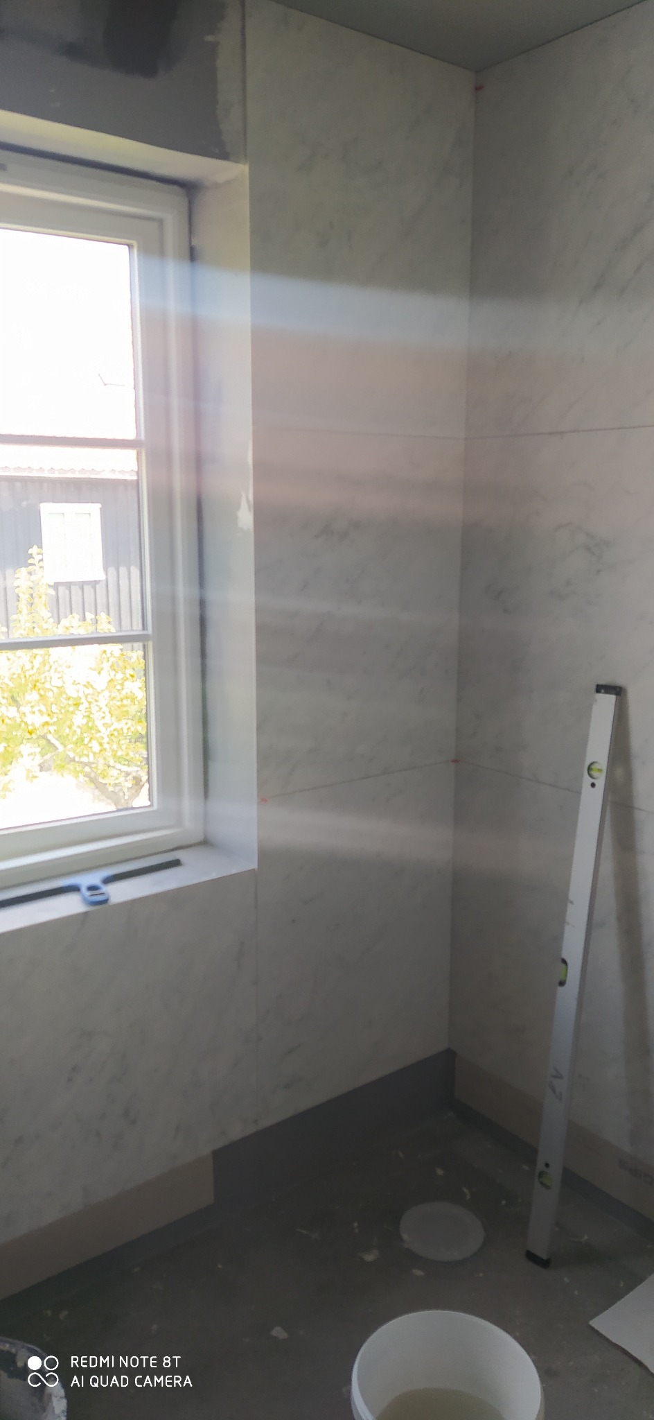 Wnętrze łazienki w trakcie prac wykończeniowych, ściany wyłożone białymi płytkami imitującymi marmur, poziomica oparta o ścianę, widoczne narzędzia i wiadro, okno z widokiem na budynek i drzewa.