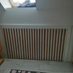 Zabudowa grzejnika z pionowymi drewnianymi lamelami w białej ramie, pod skosem dachu z oknem i leżącym ubraniem, na podłodze szary dywan w biało-różowe serca.