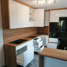 Nowoczesna kuchnia z białymi, lakierowanymi szafkami i drewnianym blatem, otwarte szuflady z systemem organizacji, oświetlenie punktowe sufitowe, widok na czarny piekarnik w zabudowie.