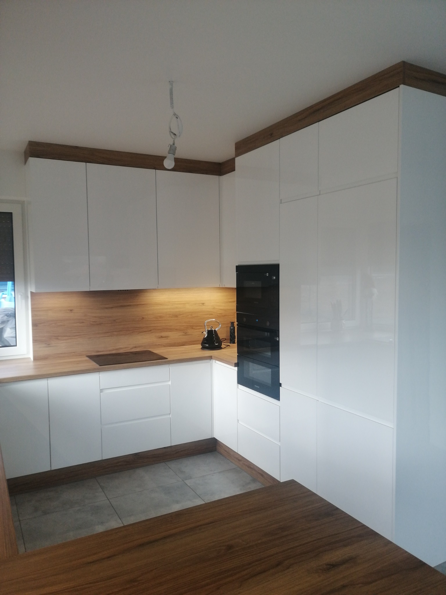 Nowoczesna kuchnia z białymi, lakierowanymi frontami, drewnianym blatem i górną listwą, widoczny fragment wyspy kuchennej z blatem w tym samym kolorze, czajnik na blacie i zabudowany piekarnik.