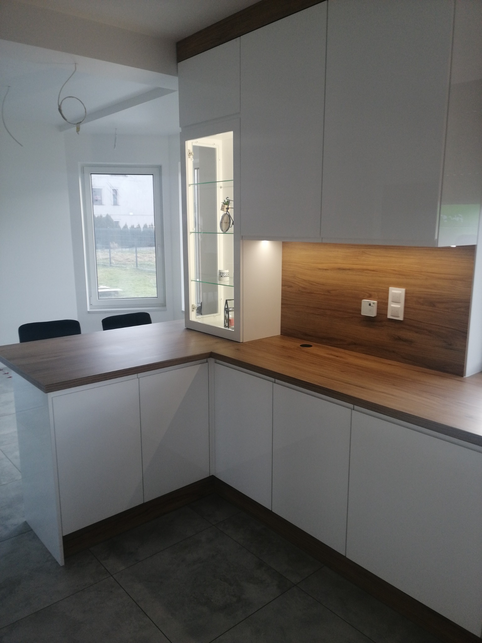 Nowoczesna kuchnia z białymi frontami i drewnianym blatem, oświetleniem LED pod szafkami wiszącymi i przeszkloną witryną narożną, widok na okno i dwa krzesła w tle.