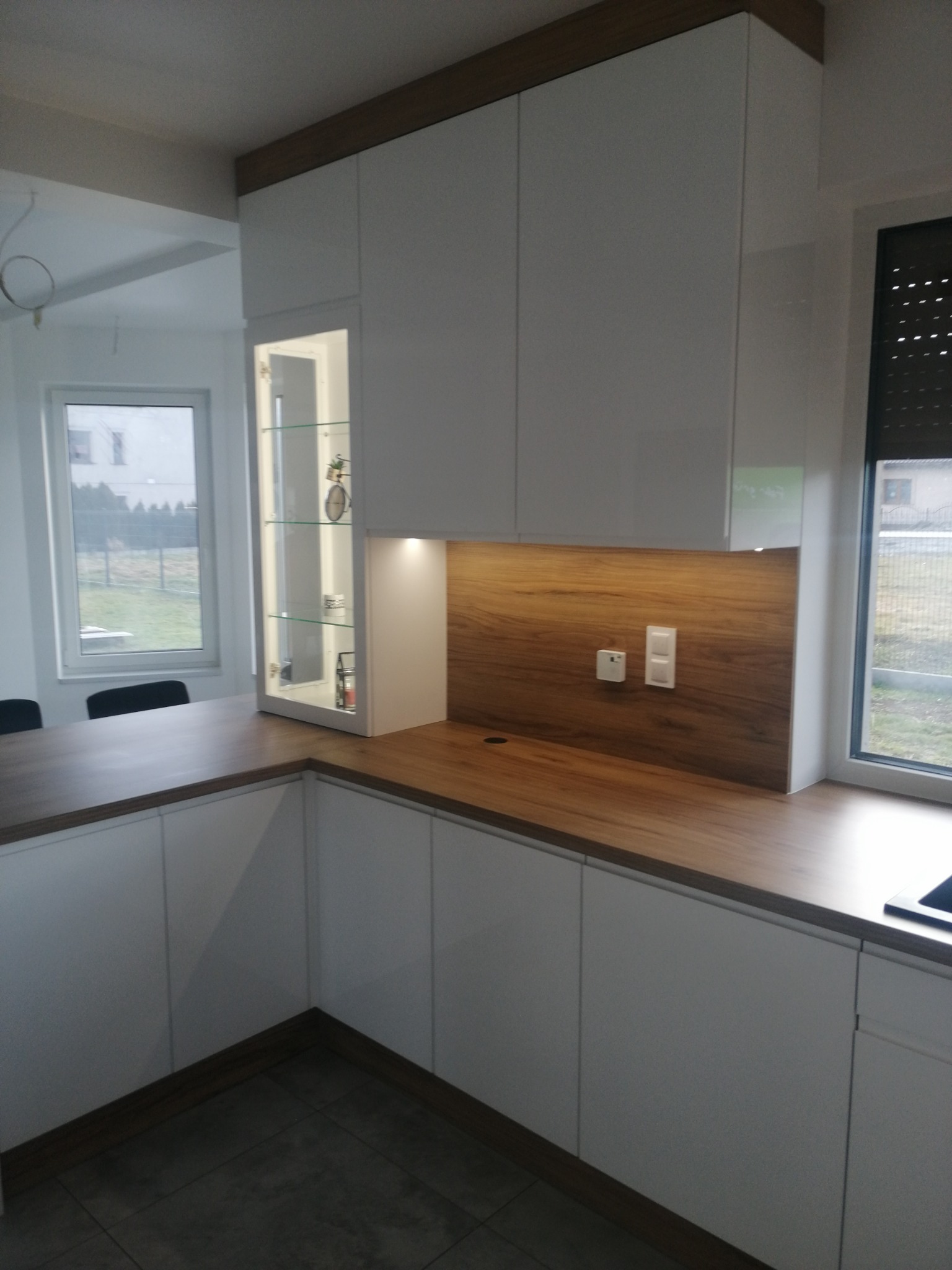 Nowoczesna biała kuchnia z drewnianym blatem i oświetloną witryną, widoczne gniazdka elektryczne na drewnianej ściance nad blatem.