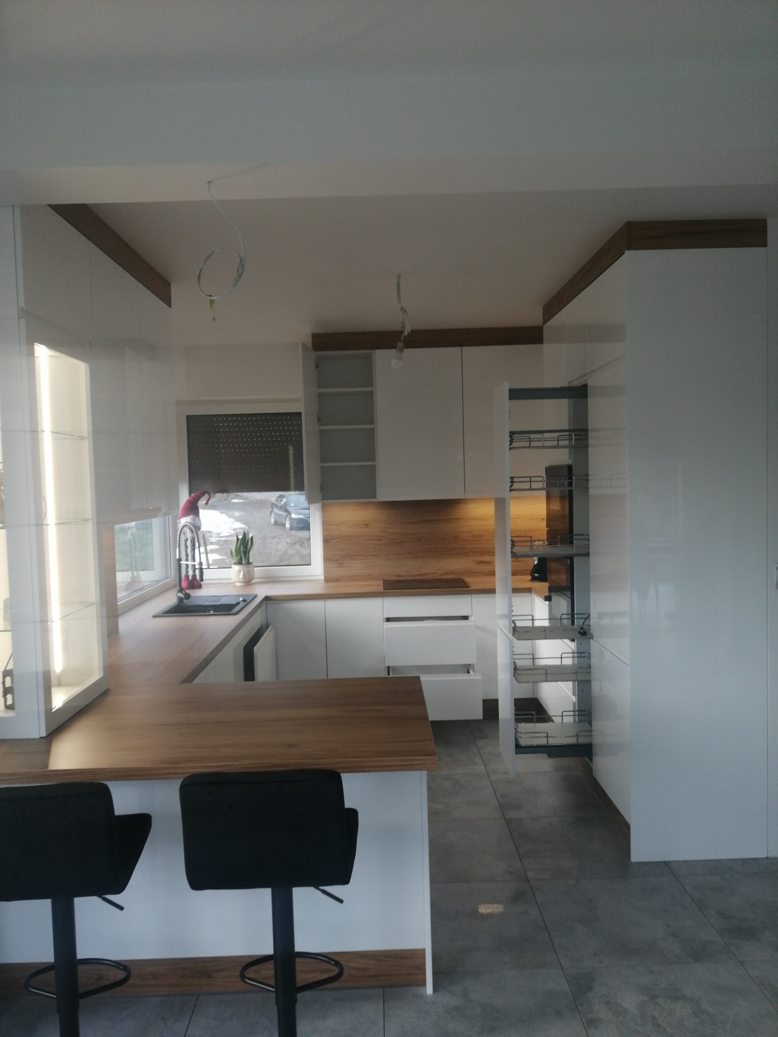 Nowoczesna kuchnia z białymi szafkami i drewnianym blatem, wysuwana szafka cargo, dwa czarne hokery przy wyspie, okno z widokiem na zewnątrz.