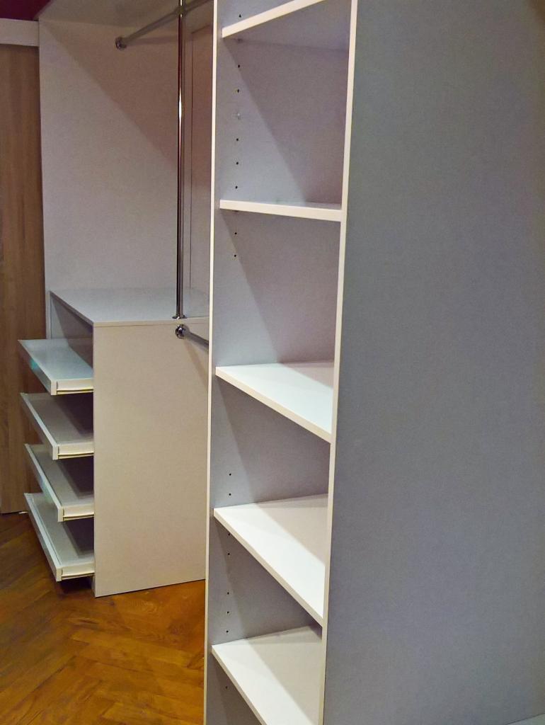 Wnętrze garderoby z wysuwanymi półkami i drążkiem na ubrania, widoczne elementy zabudowy z szarymi i białymi frontami.