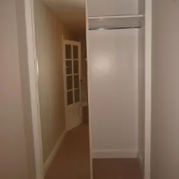 Wnęka z zabudową szafy wnękowej z przesuwnymi drzwiami lustrzanymi, widoczna pusta przestrzeń wewnątrz, z półkami i drążkiem, na podłodze wykładzina.