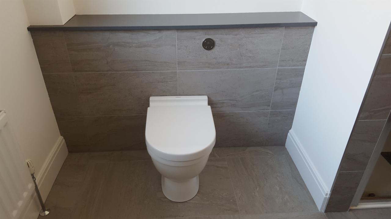 Nowoczesna toaleta podwieszana z szarymi płytkami imitującymi kamień, ukryta instalacja z przyciskiem spłukiwania w ścianie, białe listwy przypodłogowe.