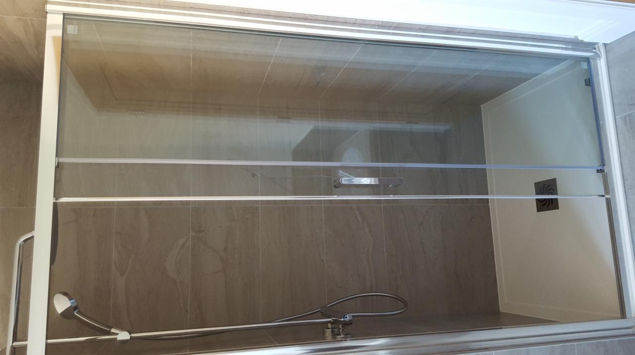 Szklana kabina prysznicowa z przesuwnymi drzwiami i chromowaną armaturą, zamontowana na ścianie z płytek imitujących kamień. Widoczny odpływ liniowy i wentylator w kabinie.