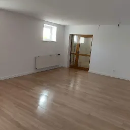 Jasne panele podłogowe w wyremontowanym pokoju z białymi ścianami, grzejnikiem pod oknem i drewnianymi drzwiami.