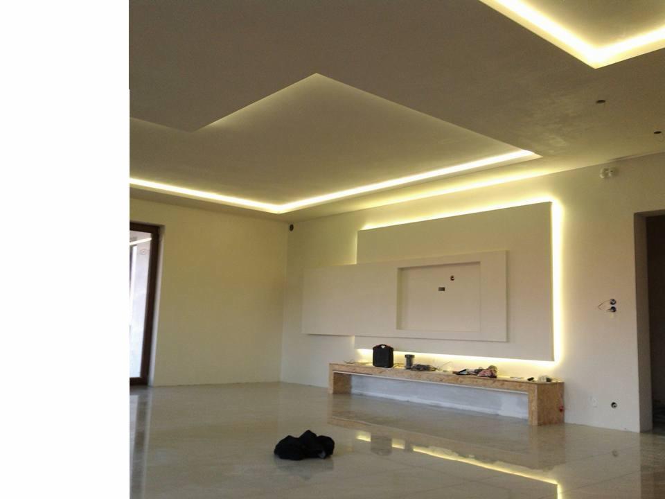 Wnętrze pokoju z podwieszanym sufitem z oświetleniem LED, białą ścianą z dekoracyjnym panelem i półką z elementami instalacji elektrycznej, widok na błyszczącą podłogę.