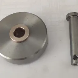 Metalowy dysk z mosiężną tuleją i metalowy trzpień z otworem, leżące na białym tle z widocznym fragmentem rysunku technicznego.