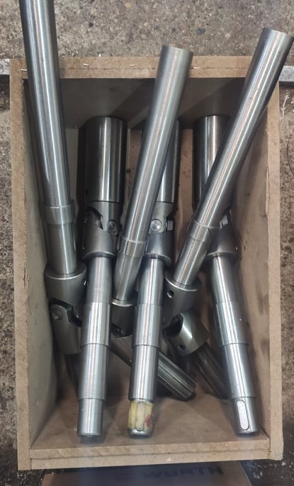 Kilkanaście metalowych wałów przegubowych w drewnianej skrzyni, widoczne ślady obróbki i montażu.