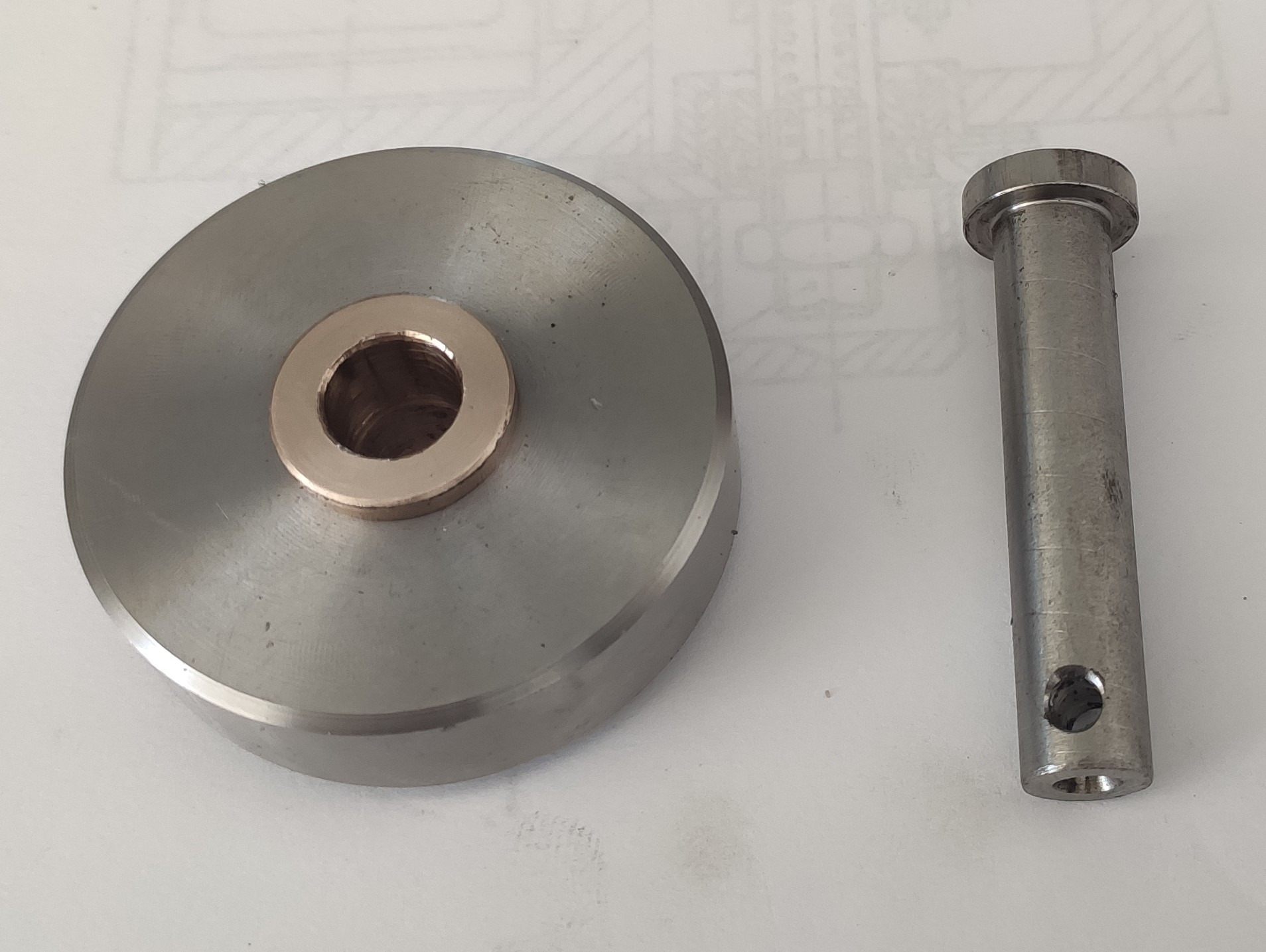 Metalowy dysk z mosiężną tuleją i metalowy trzpień z otworem, leżące na białym tle z widocznym fragmentem rysunku technicznego.