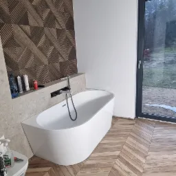 Nowoczesna łazienka z wolnostojącą wanną, drewnianą podłogą ułożoną w jodełkę i geometryczną okładziną ścienną, widok na zewnątrz przez okno.