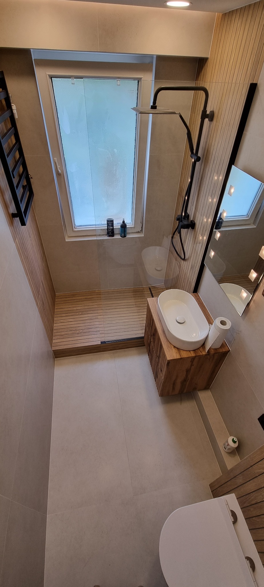 Nowoczesna łazienka na poddaszu z kabiną prysznicową typu walk-in, drewnianym wykończeniem strefy prysznica, umywalką nablatową na drewnianej szafce i oknem dachowym z widocznymi kosmetykami.
