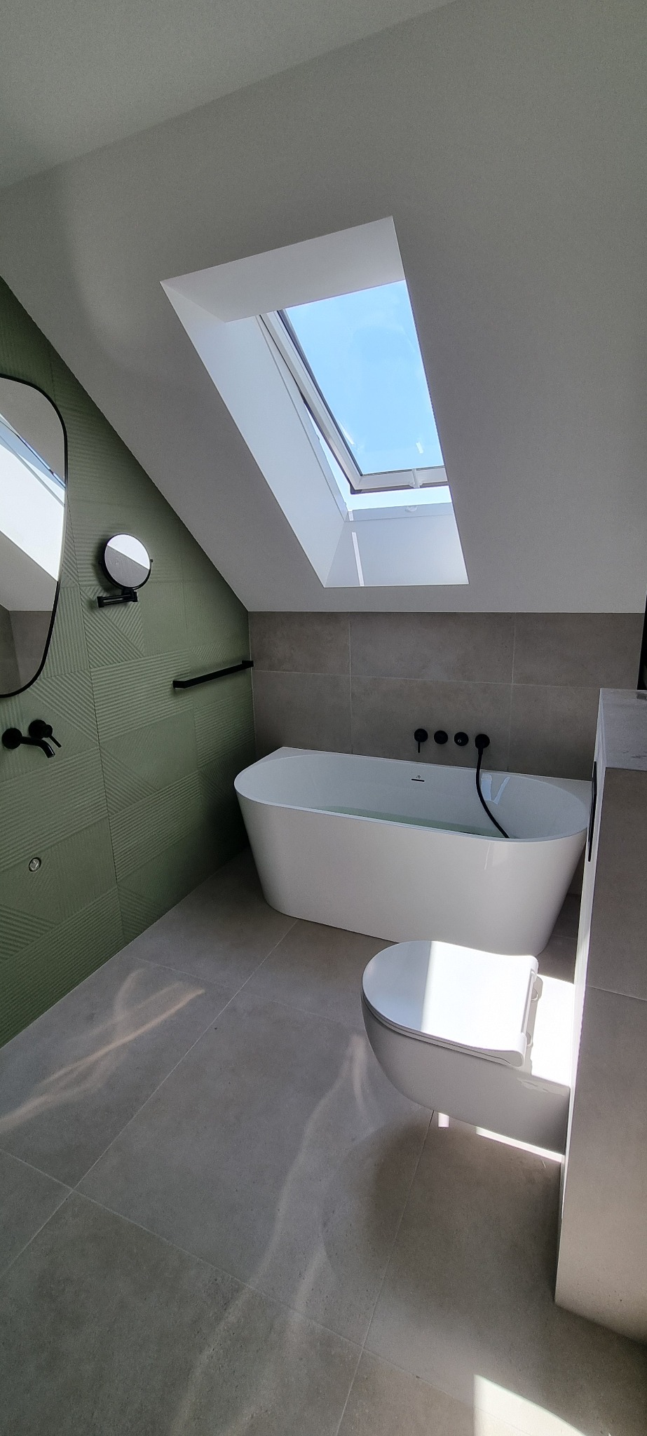 Nowoczesna łazienka na poddaszu z oknem dachowym, zieloną ścianą z geometrycznym wzorem, wolnostojącą wanną i czarną armaturą.