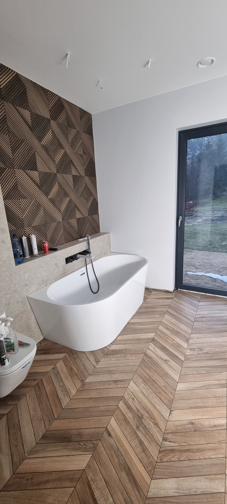 Nowoczesna łazienka z wolnostojącą wanną, drewnianą podłogą ułożoną w jodełkę i geometryczną okładziną ścienną, widok na zewnątrz przez okno.