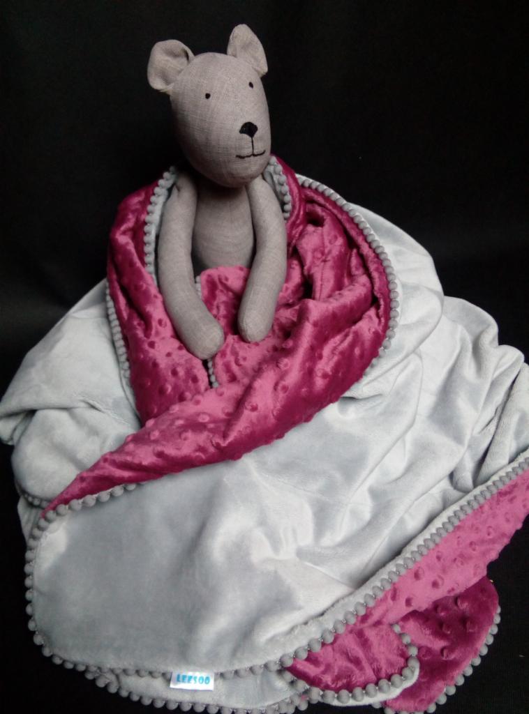 Szary pluszowy miś siedzi na miękkim, dwustronnym kocyku z szarego i fuksjowego weluru z pomponami, na czarnym tle.