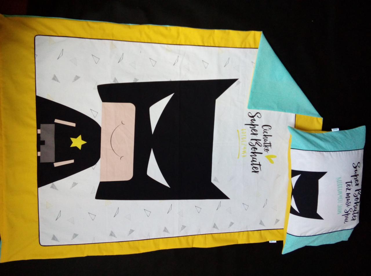 Komplet pościeli dziecięcej z motywem superbohatera, składający się z kołdry i poduszki, z żółto-turkusowymi obramowaniami na białym tle w trójkąty, leżący na czarnym tle.