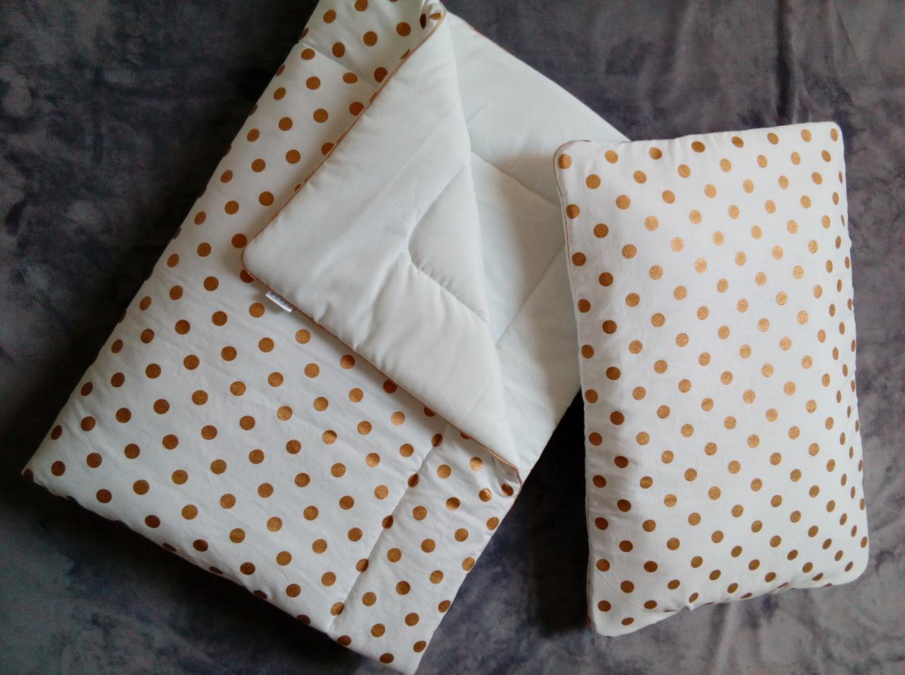 Biały śpiworek niemowlęcy i poduszka z motywem złotych kropek, leżące na szarym tle.