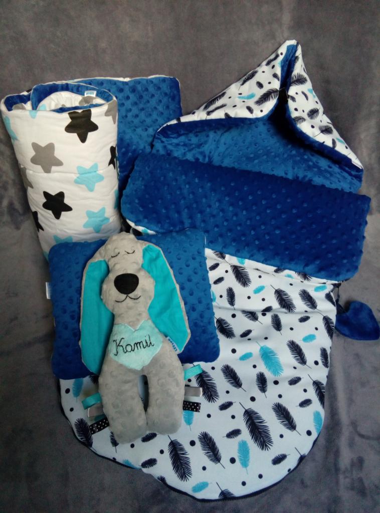 Wyprawka niemowlęca z Wrocławia: rożek, kocyk, poduszka, ochraniacz do łóżeczka i pluszowy piesek z imieniem Kamil, utrzymane w tonacji bieli, szarości i błękitu, z motywem piórek i gwiazdek.