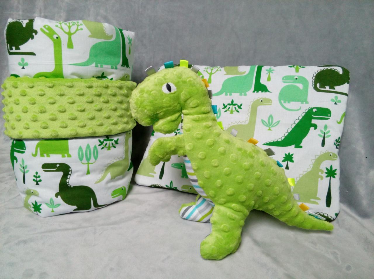 Ręcznie szyte, zielone, tekstylne pojemniki do przechowywania z motywem dinozaurów, ustawione jeden na drugim, obok poduszki z tym samym wzorem i pluszowego dinozaura.