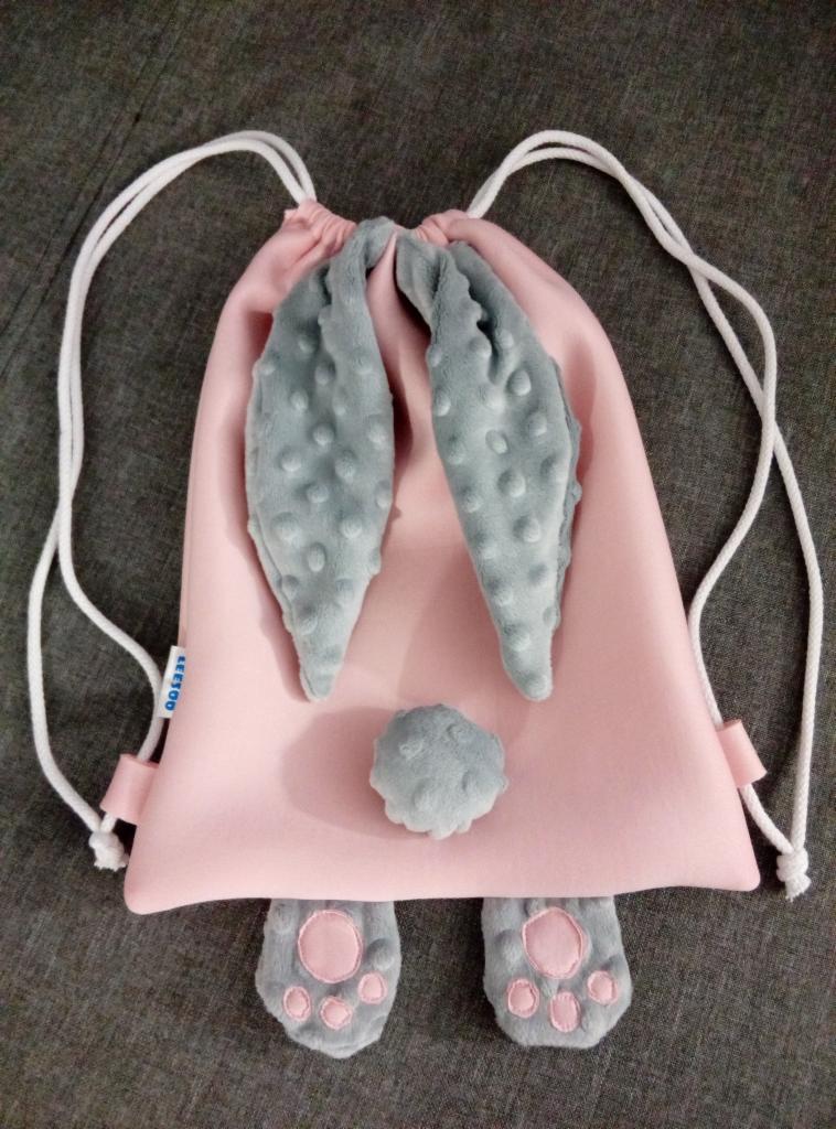 Różowy plecak-worek z aplikacjami w kształcie króliczych uszu, ogonka i łapek z szarego pluszu w kropki, leżący na szarym tle.
