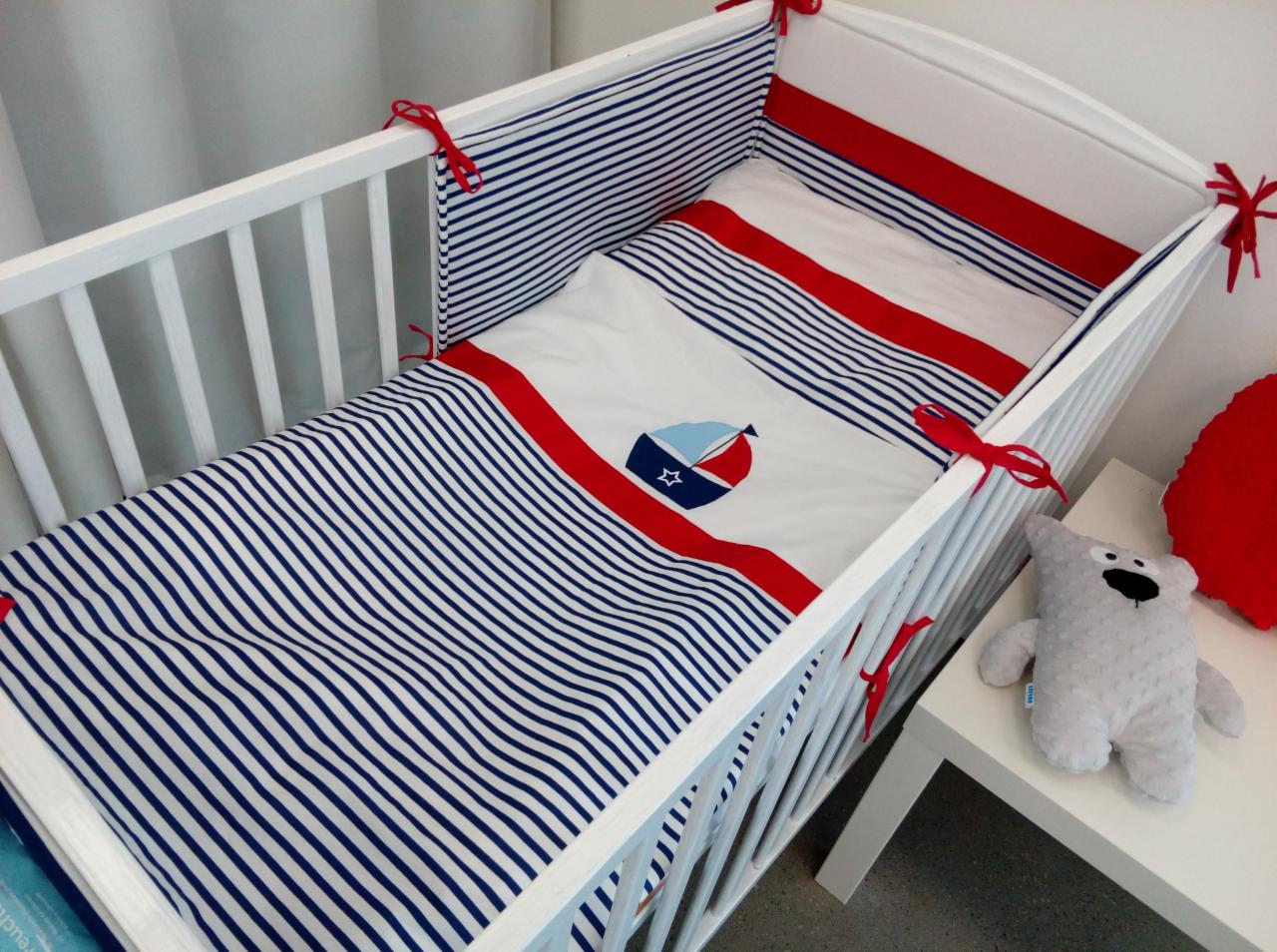 Białe łóżeczko dziecięce z pościelą w biało-niebieskie paski i czerwonymi akcentami, motyw żaglówki na kołderce, pluszowy miś na stoliku obok.