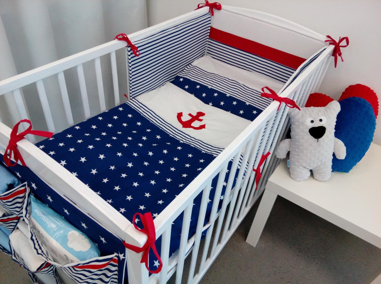 Białe łóżeczko dziecięce z marynarską pościelą w gwiazdki i kotwicę, z czerwonymi wstążkami i pluszowym misiem obok.