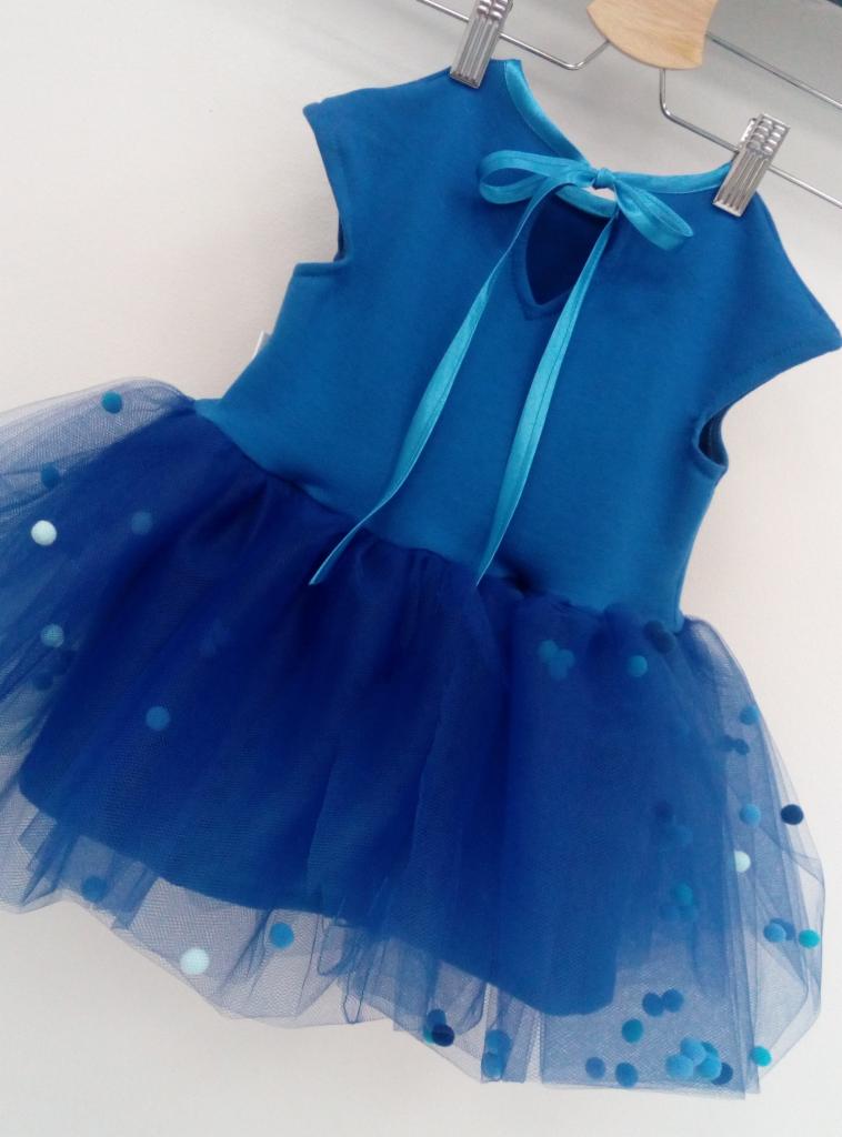 Niebieska sukienka baletowa dla dziewczynki z tiulowym dołem w kropki, wisząca na drewnianym wieszaku z metalowymi klipsami, z satynową wstążką wiązaną na szyi.