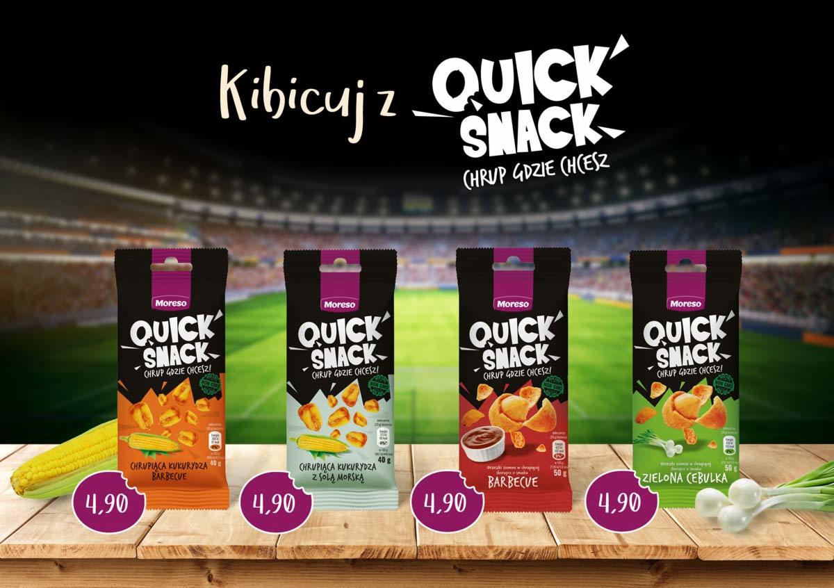 Cztery opakowania chrupiącej kukurydzy Quick Snack o różnych smakach (barbecue, z solą morską, barbecue, zielona cebulka) ustawione na drewnianym stole z cenami 4,90 zł, w tle stadion piłkarski.