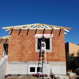 SDG-BAU KG - Montaż okna w surowym budynku z czerwonej cegły, pracownik na drabinie umieszcza piankę izolacyjną wokół ramy okiennej, widoczne drewniane elementy konstrukcji dachu.