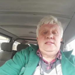 Mężczyzna w zielonej kurtce siedzący w pojeździe, prawdopodobnie w trakcie przerwy w pracy. Widoczny fragment wnętrza samochodu i ubranie robocze.