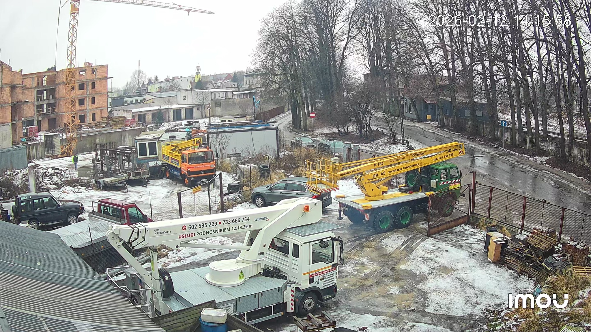Dwa podnośniki koszowe na placu budowy w zimowej scenerii. Jeden biały z wysięgnikiem, drugi żółto-zielony. W tle budynek w trakcie budowy i drzewa.