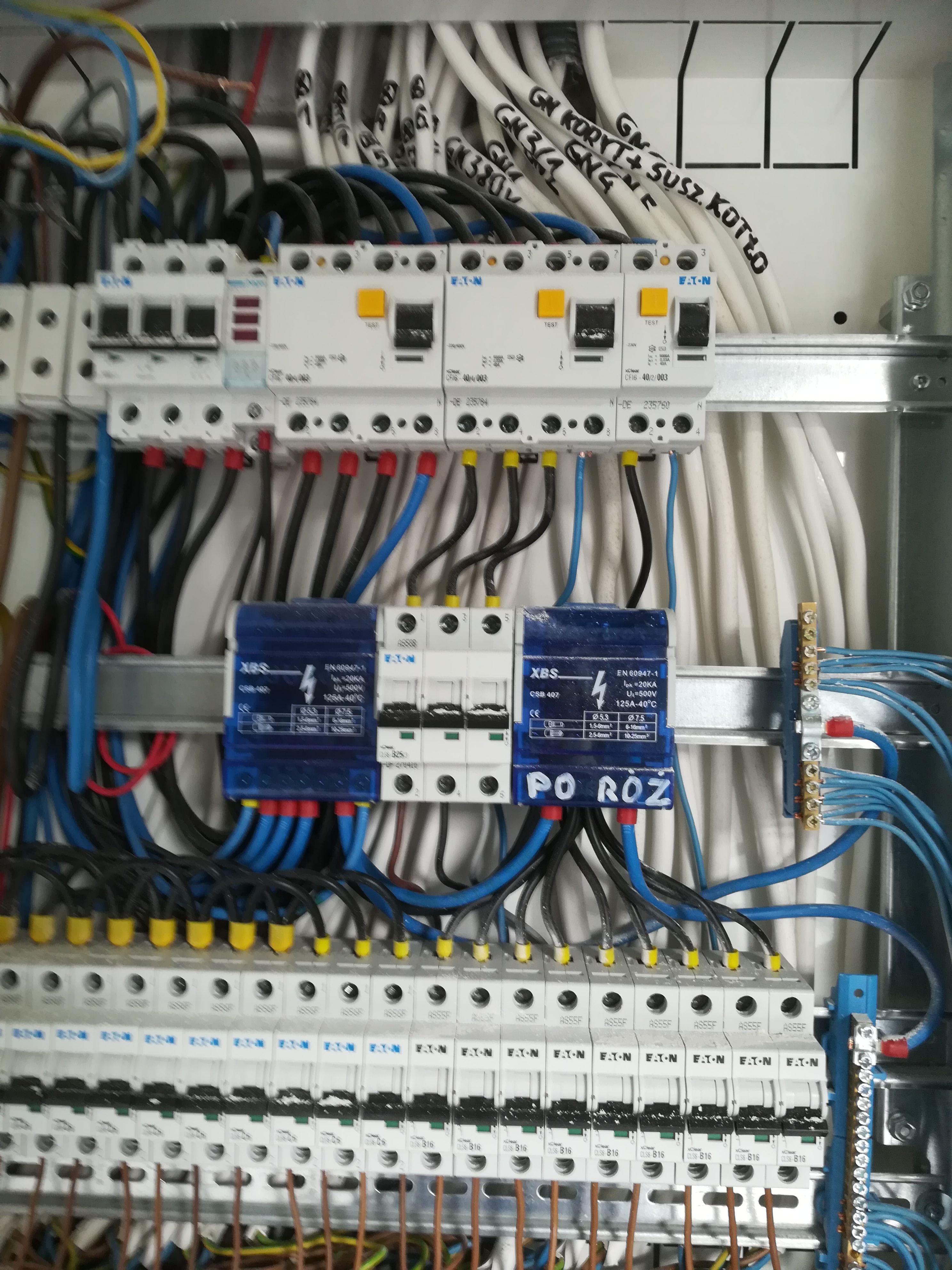 Rozdzielnica elektryczna z zainstalowanymi bezpiecznikami i okablowaniem, widoczne oznaczenia przewodów i komponentów, w tym napisy 'PO ROZ' na jednym z elementów.